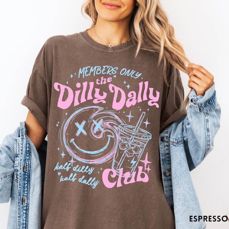 May include: Espresso-colored t-shirt with a graphic design. The design features the text "MEMBERS ONLY the Dilly Dally Club" in pink and blue, along with a smiley face and a beverage illustration. The shirt has rolled-up sleeves.