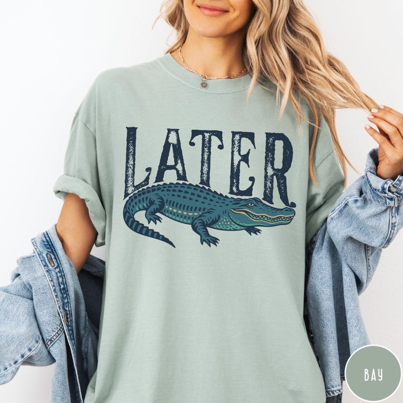 May include: A light green t-shirt with the word "LATER" in dark blue above a blue alligator illustration. The alligator has yellow accents and is facing right. The shirt has rolled-up sleeves.