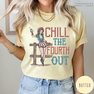 May include: Butter-colored t-shirt with a graphic featuring a pin-up girl in patriotic attire, holding a firecracker. The text "CHILL THE FOURTH OUT" is displayed in red and blue. The design includes a wooden fence.