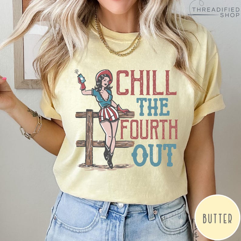 May include: Butter-colored t-shirt with a graphic featuring a pin-up girl in patriotic attire, holding a firecracker. The text "CHILL THE FOURTH OUT" is displayed in red and blue. The design includes a wooden fence.