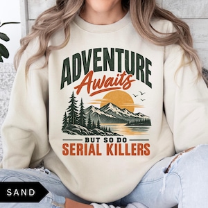 May include: A sand-colored crewneck sweatshirt featuring a graphic print of a mountain landscape with pine trees, a sunset, and birds. The text reads ADVENTURE Awaits BUT SO DO SERIAL KILLERS in dark green and orange lettering across the chest.