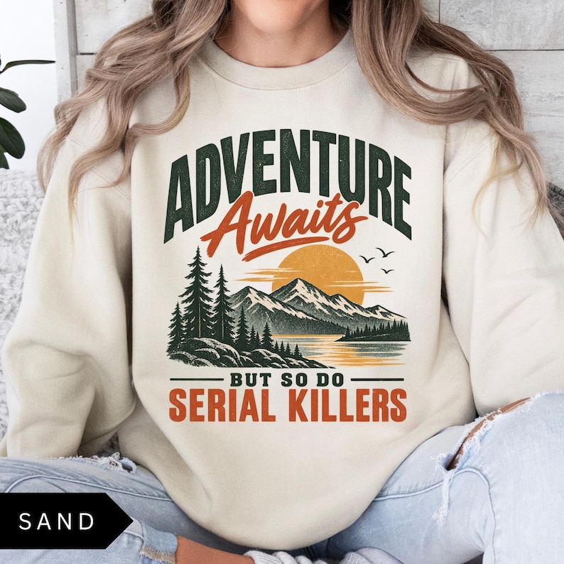 May include: A sand-colored crewneck sweatshirt featuring a graphic print of a mountain landscape with pine trees, a sunset, and birds. The text reads ADVENTURE Awaits BUT SO DO SERIAL KILLERS in dark green and orange lettering across the chest.