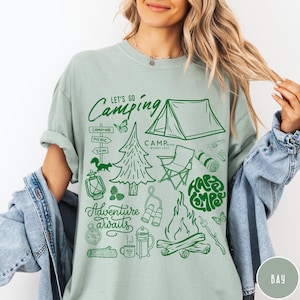 Może przedstawiać: Jasnozielona koszulka z zieloną grafiką o tematyce kempingowej. Projekt zawiera namiot, ognisko, drzewa oraz napisy "Let's Go Camping", "Adventure Awaits" i "Happy Camper".