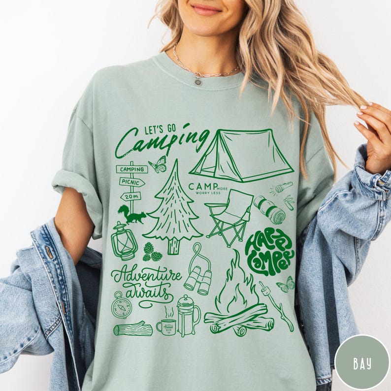 Może przedstawiać: Jasnozielona koszulka z zieloną grafiką o tematyce kempingowej. Projekt zawiera namiot, ognisko, drzewa oraz napisy "Let's Go Camping", "Adventure Awaits" i "Happy Camper".