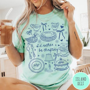 May include: A pale green t-shirt with a blue illustration of vintage kitchenware and the text "I'd rather be thrifting." The design includes plates, teacups, a duck, and a lamp.