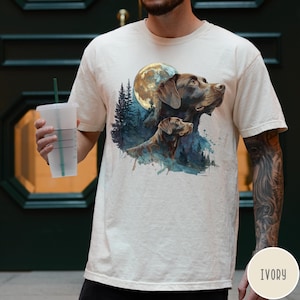 May include: Ivory-colored t-shirt featuring a watercolor illustration of two brown dogs with a full moon and forest background. The dogs are looking upwards. The shirt is a neutral color.