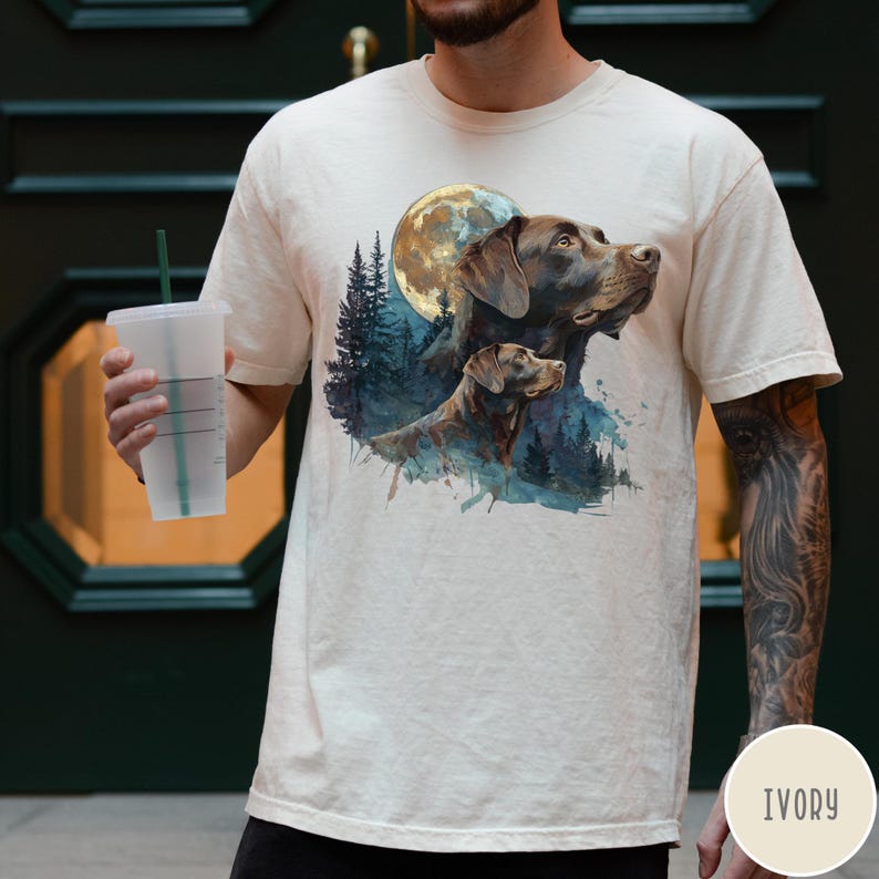 May include: Ivory-colored t-shirt featuring a watercolor illustration of two brown dogs with a full moon and forest background. The dogs are looking upwards. The shirt is a neutral color.