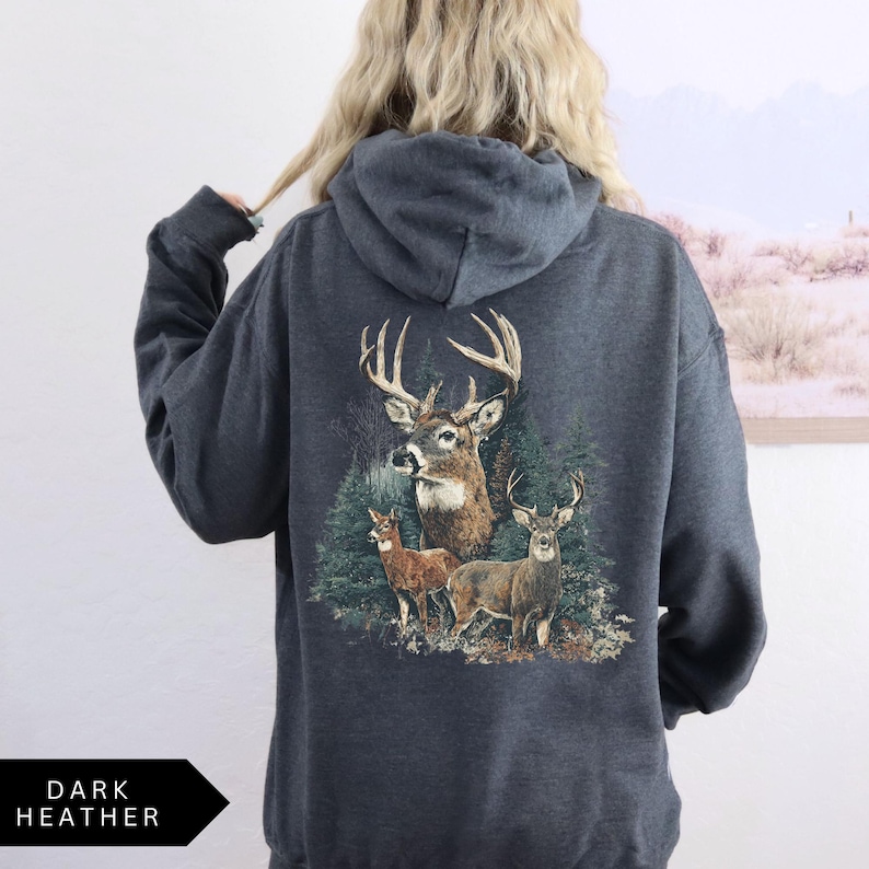 Op de afbeelding: Donkergrijze hoodie met een gedetailleerde illustratie van herten in een bos. De afbeelding toont een bok met grote geweien en twee kleinere herten. De tekst "DARK HEATHER" staat onderaan.