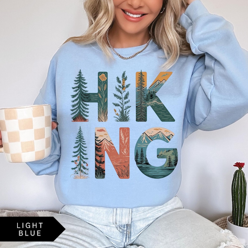 Puede incluir: Sudadera azul claro con la palabra "HIKING" en letras grandes y coloridas. Las letras est&aacute;n dise&ntilde;adas con ilustraciones de tem&aacute;tica natural de &aacute;rboles, monta&ntilde;as y plantas. Las palabras "LIGHT BLUE" est&aacute;n impresas en un rect&aacute;ngulo negro en la parte inferior.