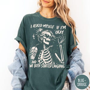 May include: Dark teal t-shirt with a skeleton graphic wearing sunglasses and holding a drink. The text reads, "I ASKED MYSELF IF I'M OKAY. AND WE BOTH STARTED LAUGHING." The shirt has rolled-up sleeves.