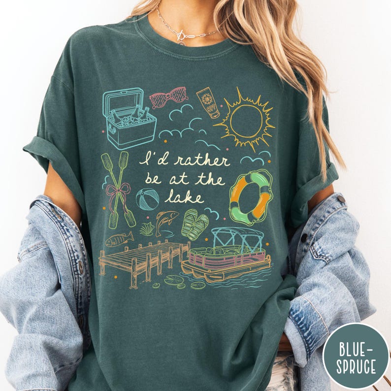 Puede incluir: Camiseta verde oscuro con un dise&ntilde;o gr&aacute;fico que presenta ilustraciones con tem&aacute;tica de lago. El dise&ntilde;o incluye una nevera, gafas de sol, protector solar, un sol, nubes, remos, una pelota de playa, un pez, sandalias, un salvavidas, un muelle y una lancha pont&oacute;n. El texto dice "I'd rather be at the lake."