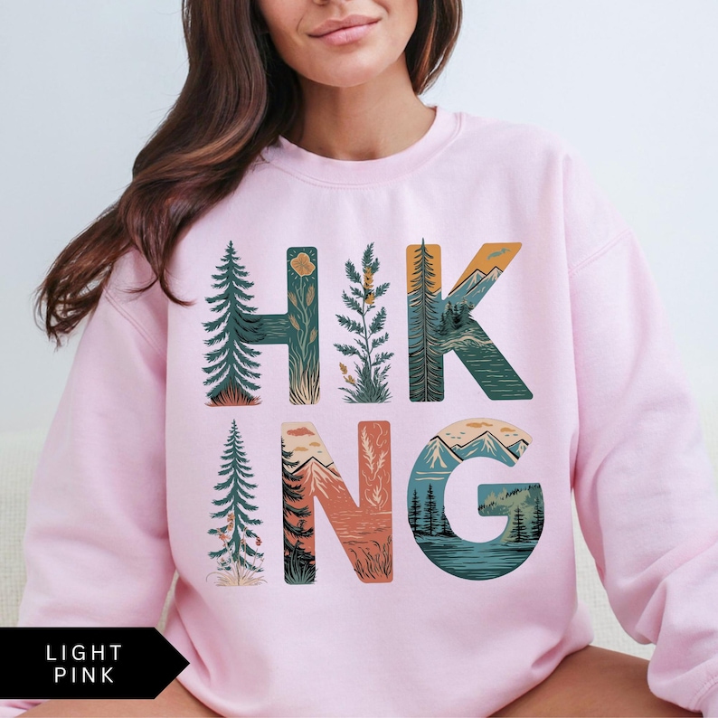 Puede incluir: Sudadera rosa claro con la palabra "HIKING" en un dise&ntilde;o tem&aacute;tico de la naturaleza. Las letras est&aacute;n rellenas de ilustraciones de &aacute;rboles, monta&ntilde;as y paisajes. La esquina inferior izquierda tiene el texto "LIGHT PINK" en una flecha negra.