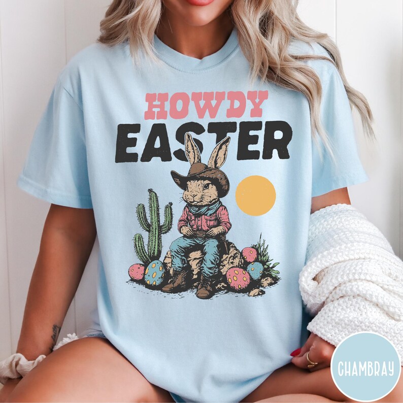 Puede incluir: Camiseta azul claro con las palabras "HOWDY EASTER" en rosa y negro. El gr&aacute;fico presenta un conejo con sombrero de vaquero sentado en una roca con huevos de Pascua y un cactus. Tambi&eacute;n hay un c&iacute;rculo amarillo.