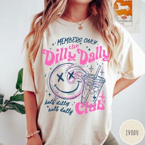 May include: Ivory-colored t-shirt with a graphic design. The design features the text "MEMBERS ONLY the Dilly Dally Club" in pink, along with a smiley face and a drink. The phrase "half dilly half dally" is also included.