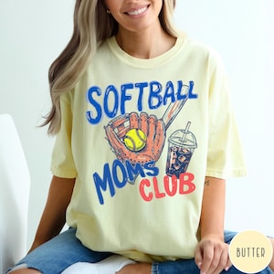 May include: Butter yellow t-shirt with the words "SOFTBALL MOMS CLUB" in blue and red. The graphic includes a softball glove, a yellow softball, and a drink with ice. The shirt is a casual, short-sleeved style.