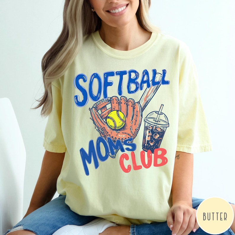 May include: Butter yellow t-shirt with the words "SOFTBALL MOMS CLUB" in blue and red. The graphic includes a softball glove, a yellow softball, and a drink with ice. The shirt is a casual, short-sleeved style.