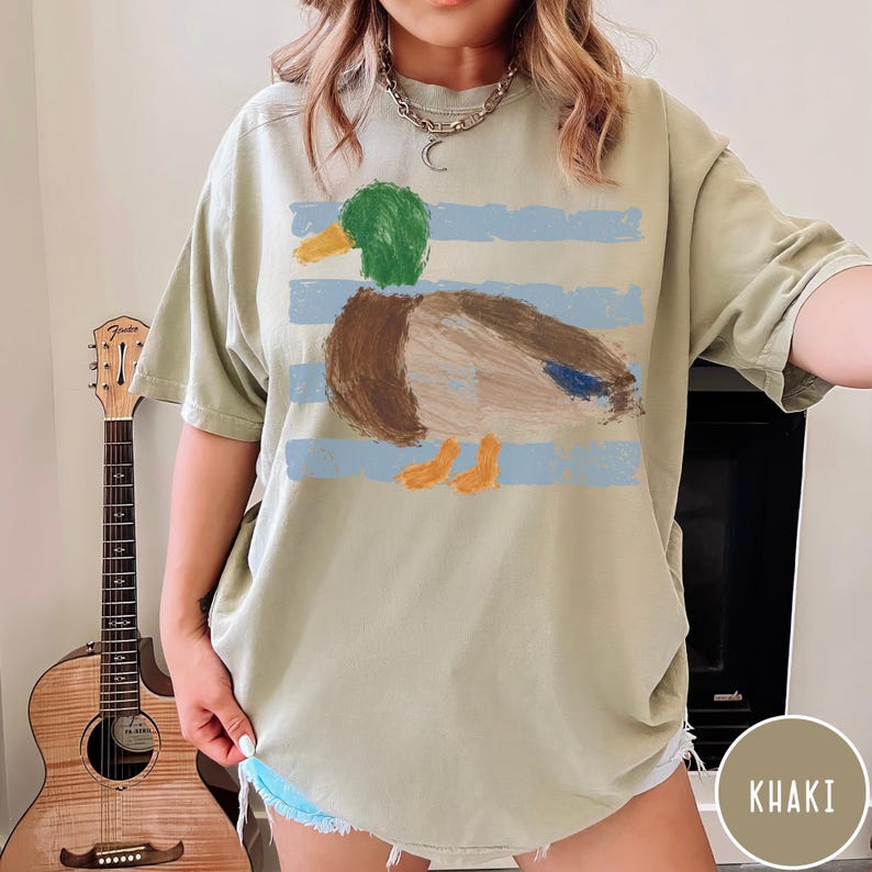 May include: A khaki-colored short-sleeve t-shirt featuring a graphic print of a mallard duck with a green head, brown body, and orange feet. The duck is layered over horizontal light blue brushstroke stripes. The shirt has a crew neckline.