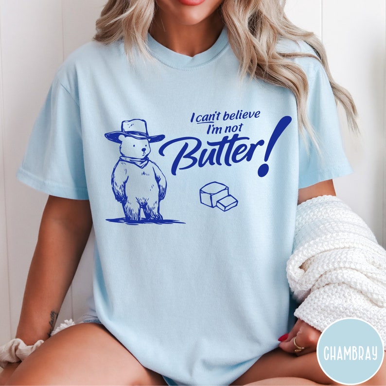 May include: A light blue t-shirt with a cartoon bear wearing a cowboy hat and the text "I can't believe I'm not Butter!" in blue.