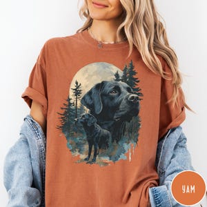 May include: Burnt orange t-shirt featuring a graphic of two black Labrador Retrievers in a forest setting with a full moon. The design includes pine trees and a watercolor effect. The shirt has short sleeves and a relaxed fit.