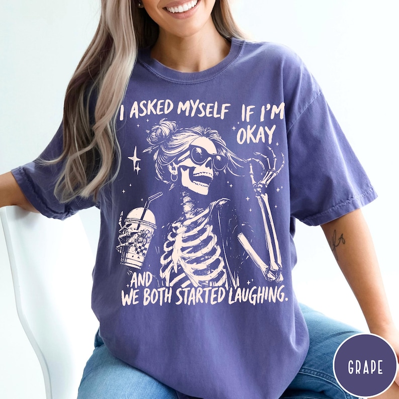 May include: A purple t-shirt with a skeleton graphic wearing sunglasses and holding a drink. The text on the shirt reads, "I asked myself if I'm okay, and we both started laughing."