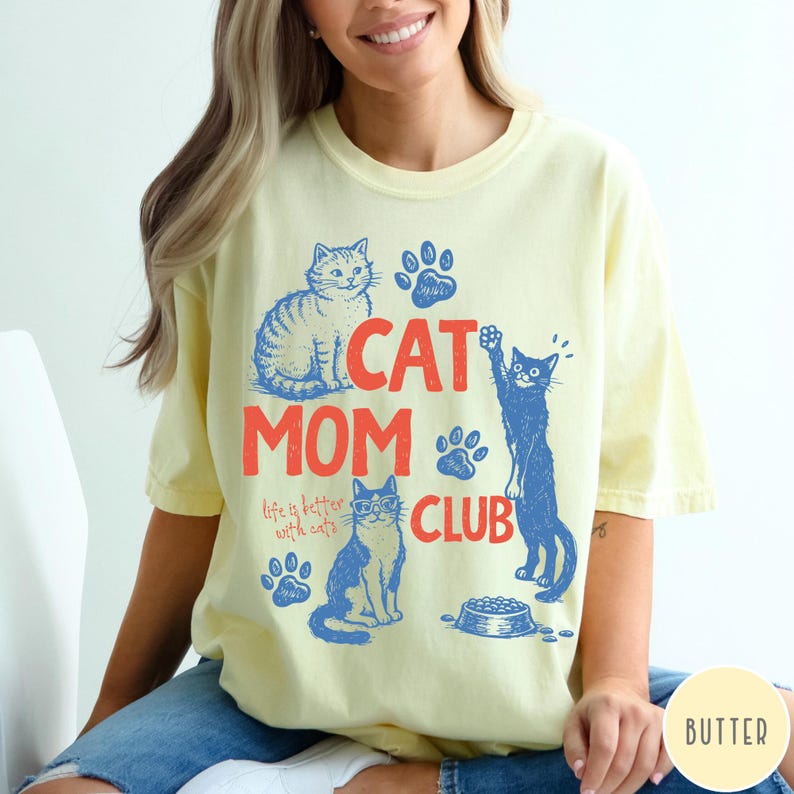 K&ouml;nnte beinhalten: Hellgelbes T-Shirt mit einer blau-orangenen Grafik. Die Grafik enth&auml;lt den Text "CAT MOM CLUB" sowie Illustrationen von Katzen, Pfotenabdr&uuml;cken und einem Futternapf. Der Text "life is better with cats" ist ebenfalls enthalten.