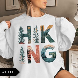 Puede incluir: Sudadera blanca con la palabra "HIKING" en letras coloridas con tem&aacute;tica de la naturaleza. Las letras est&aacute;n dise&ntilde;adas con im&aacute;genes de &aacute;rboles, monta&ntilde;as y paisajes. La palabra "WHITE" est&aacute; en un rect&aacute;ngulo negro en la parte inferior.