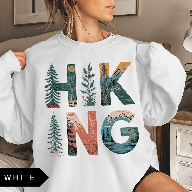 Puede incluir: Sudadera blanca con la palabra "HIKING" en letras coloridas con tem&aacute;tica de la naturaleza. Las letras est&aacute;n dise&ntilde;adas con im&aacute;genes de &aacute;rboles, monta&ntilde;as y paisajes. La palabra "WHITE" est&aacute; en un rect&aacute;ngulo negro en la parte inferior.
