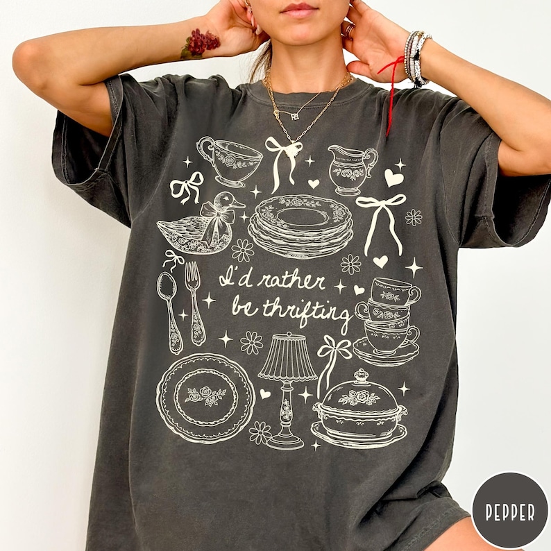 May include: Dark grey t-shirt featuring a white graphic of kitchenware, such as plates, cups, and a duck. The text "I'd rather be thrifting" is printed on the shirt. A casual, vintage-style tee.
