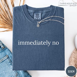 May include: A denim blue t-shirt with the words "immediately no" printed in white. A silver necklace with a sunburst pendant is draped across the shirt. The shirt is folded and has a Comfort Colors tag.