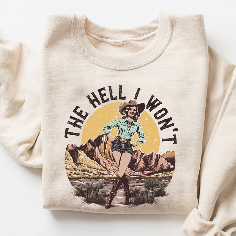 May include: A beige sweatshirt with a vintage-style graphic print of a woman wearing a cowboy hat and boots. The graphic says "The Hell I Won't".