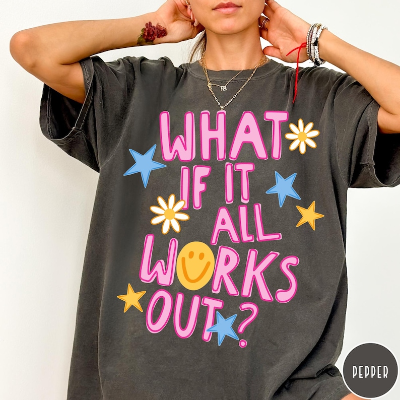 May include: A charcoal gray short-sleeve t-shirt featuring the pink text WHAT IF IT ALL WORKS OUT? along with yellow smiley face, white daisy, and blue and yellow star graphics. The shirt has a crew neckline and a relaxed, loose-fitting silhouette.