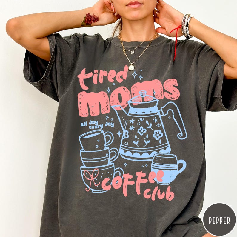 May include: Dark gray t-shirt with the words "tired moms coffee club" in pink and blue. The design includes a teapot, stacked mugs, and the phrase "all day every day". The shirt has a vintage, worn look.