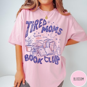 May include: Pink t-shirt with the text "Tired Moms Book Club" in blue script. The design includes illustrations of books, candles, plants, and stars. The shirt also has the text "Running on coffee, chaos & chapters."