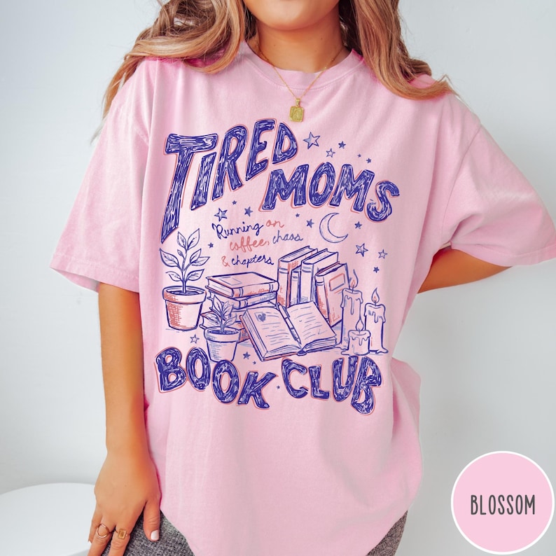 May include: Pink t-shirt with the text "Tired Moms Book Club" in blue script. The design includes illustrations of books, candles, plants, and stars. The shirt also has the text "Running on coffee, chaos & chapters."