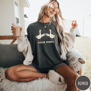 Puede incluir: Una camiseta de cuello redondo en color gris carb&oacute;n con un gr&aacute;fico de dos gansos blancos chocando los pu&ntilde;os. Debajo de los gansos, el texto GOOSE BUMPS aparece en una fuente blanca sin remates. La camiseta tiene un corte holgado y casual.