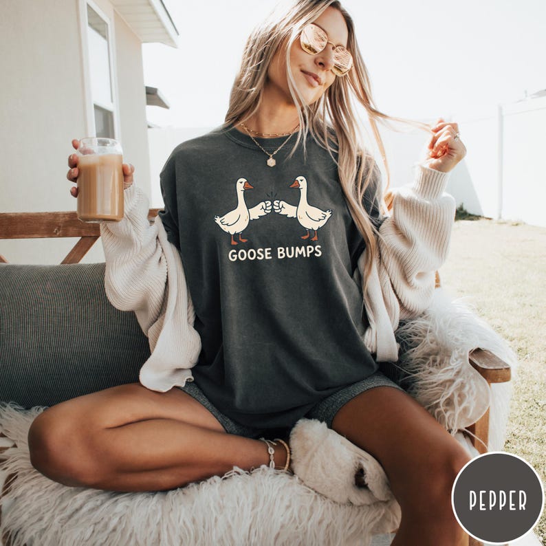 Puede incluir: Una camiseta de cuello redondo en color gris carb&oacute;n con un gr&aacute;fico de dos gansos blancos chocando los pu&ntilde;os. Debajo de los gansos, el texto GOOSE BUMPS aparece en una fuente blanca sin remates. La camiseta tiene un corte holgado y casual.