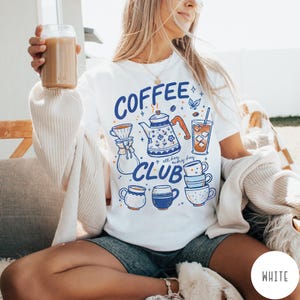 May include: White t-shirt with a blue and orange graphic that says "COFFEE CLUB" with coffee-related illustrations. The design includes a teapot, coffee cups, and a glass of iced coffee. The person is holding a glass of coffee.