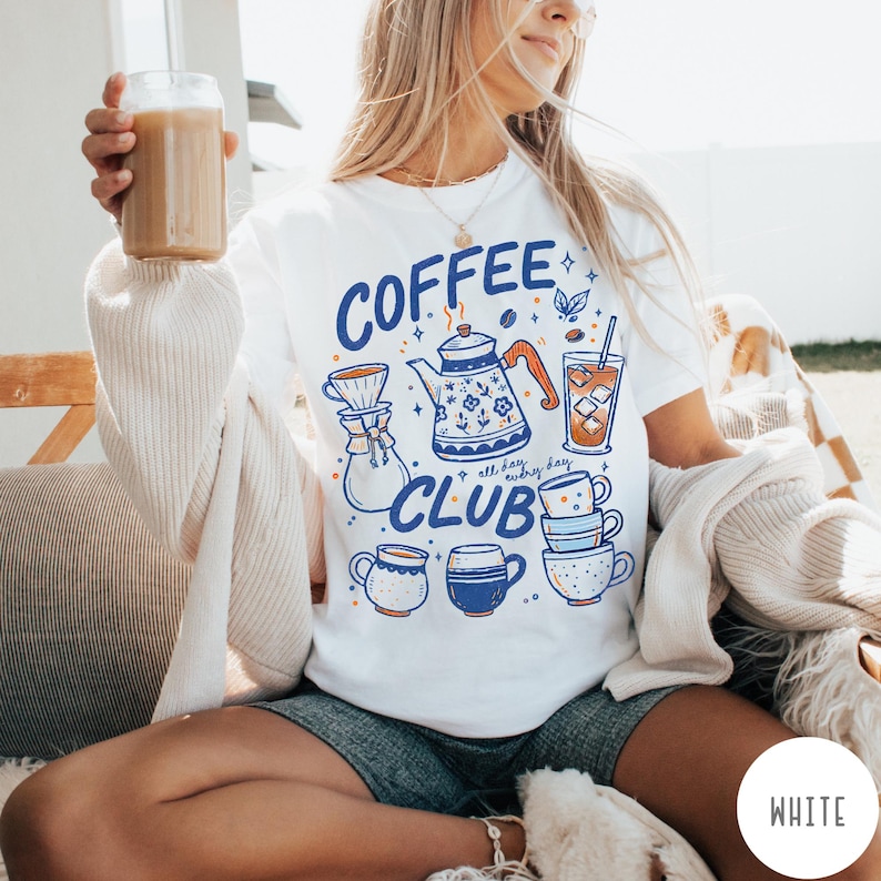 May include: White t-shirt with a blue and orange graphic that says "COFFEE CLUB" with coffee-related illustrations. The design includes a teapot, coffee cups, and a glass of iced coffee. The person is holding a glass of coffee.
