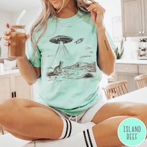 Pode incluir: Uma mulher vestindo uma camiseta verde claro com uma ilustra&ccedil;&atilde;o em preto e branco de um gato olhando para cima para um OVNI. O OVNI est&aacute; emitindo raios de luz. O texto "Island Reef" &eacute; vis&iacute;vel no canto inferior direito da imagem.