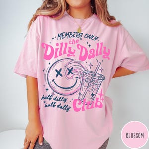 May include: Pink t-shirt with a graphic design featuring a smiling face with crossed-out eyes, a drink with ice, and the text "MEMBERS ONLY the Dilly Dally Club." The shirt also has the text "half dilly half dally."
