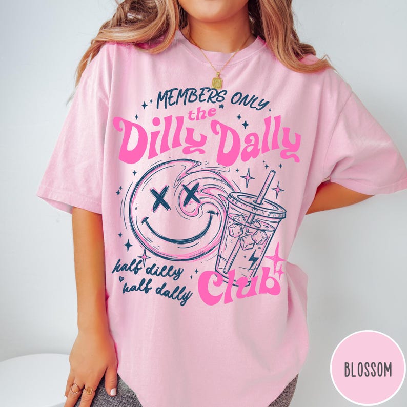 May include: Pink t-shirt with a graphic design featuring a smiling face with crossed-out eyes, a drink with ice, and the text "MEMBERS ONLY the Dilly Dally Club." The shirt also has the text "half dilly half dally."