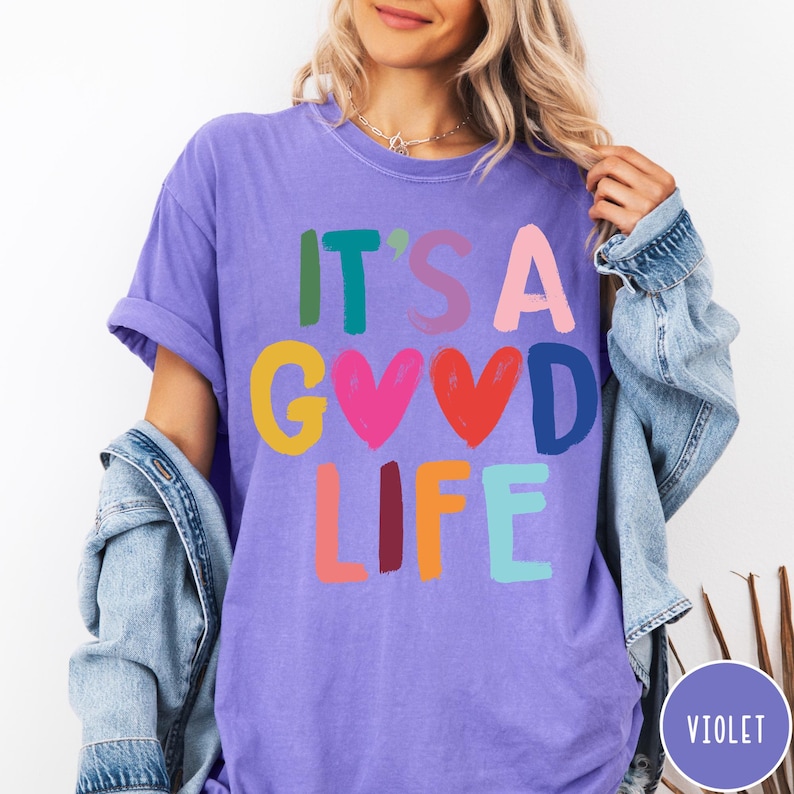 K&ouml;nnte beinhalten: Ein violettes Kurzarm-T-Shirt mit dem mehrfarbigen Schriftzug IT'S A GOOD LIFE, bei dem zwei Herzformen den Buchstaben O in GOOD ersetzen. Das T-Shirt hat einen Rundhalsausschnitt und wird unter einer blauen Jeansjacke mit hochgekrempelten &Auml;rmeln getragen.