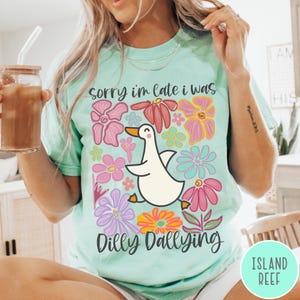 May include: A light blue t-shirt with a white duck and colorful flowers. The text "sorry i'm late i was dilly dallying" is printed on the shirt.