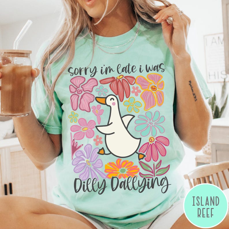 May include: A light blue t-shirt with a white duck and colorful flowers. The text "sorry i'm late i was dilly dallying" is printed on the shirt.