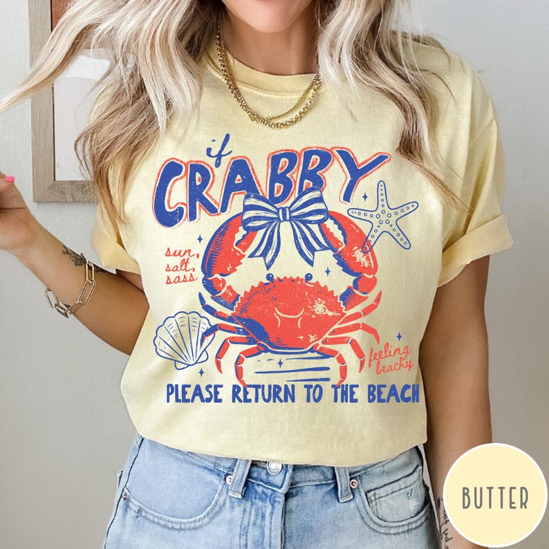 Pode incluir: Camiseta amarelo-claro com um desenho de caranguejo vermelho e azul e o texto "if Crabby, Please Return to the Beach". O design inclui uma concha e uma estrela do mar, com as palavras "sun, salt, sass" e "feeling beachy".
