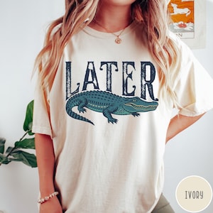 May include: Ivory-colored t-shirt featuring the word "LATER" in dark blue, above a teal alligator illustration. The alligator has a detailed design and is facing right. The shirt is a casual style.