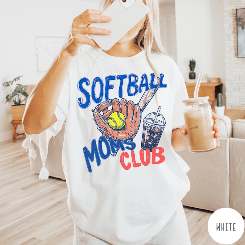 May include: White t-shirt with a graphic design featuring a softball glove, a yellow softball, and a drink with ice. The text on the shirt reads "SOFTBALL MOMS CLUB" in blue and red.