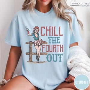 May include: Light blue t-shirt with a graphic of a pin-up girl holding a firework, standing by a wooden fence. The text "CHILL THE FOURTH OUT" is in red and blue. The shirt is a relaxed fit.