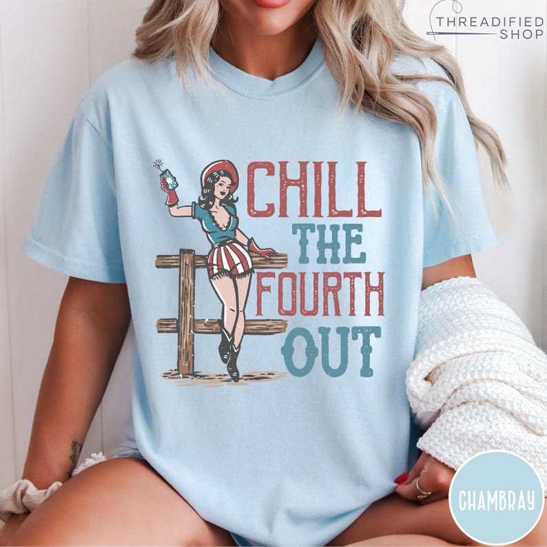 May include: Light blue t-shirt with a graphic of a pin-up girl holding a firework, standing by a wooden fence. The text "CHILL THE FOURTH OUT" is in red and blue. The shirt is a relaxed fit.
