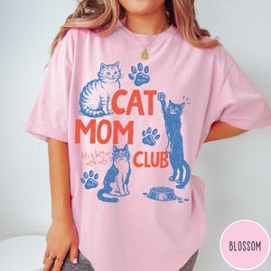 K&ouml;nnte beinhalten: Hellrosa T-Shirt mit blau-orangem Aufdruck. Der Aufdruck zeigt "CAT MOM CLUB" mit Katzen-Illustrationen und Pfotenabdr&uuml;cken. Der Text "life is better with cats" ist ebenfalls enthalten.